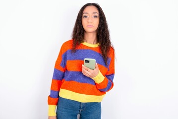 Upset dissatisfied beautiful teen girl wearing knitted colourful sweater over white background uses mobile software application and surfs information in internet, holds modern mobile hand