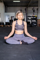 Smiley and relaxed mid adult woman resting and meditating in easy pose (sukhasana) with her eyes closed during her vinyasa flow yoga practice alone wearing sportswear indoors