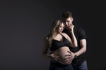 A young expectant couple looking down at their baby while the father is holding her belly in a studio on a dark background: Edmonton, Alberta, Canada