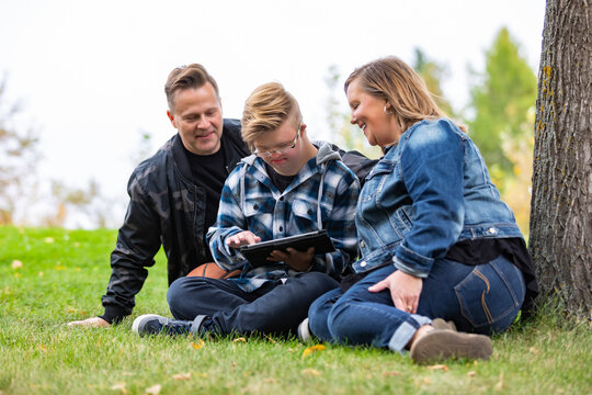 A young man with Down Syndrome learns a new program on a tablet with his father and mother while enjoying each other's company in a city park on a warm fall evening; Edmonton, Alberta, Canada