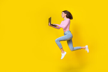 Full length photo of cheerful cute girl wear striped top denim jeans run writing message on laptop isolated on yellow color background