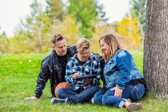 A young man with Down Syndrome learns a new program on a tablet with his father and mother while enjoying each other's company in a city park on a warm fall evening; Edmonton, Alberta, Canada
