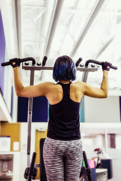 A view from behind of a paraplegic women doing pull-ups to increase her upper body strength, in a fitness facility: Sherwood Park, Alberta, Canada