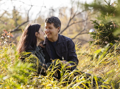 Portrait Of Mature Couple Sitting In The Tall Grass Looking At Each Other Lovingly And Enjoying Each Other’s Company While On A Walk In The Woods In Autumn; St Albert, Alberta, Canada