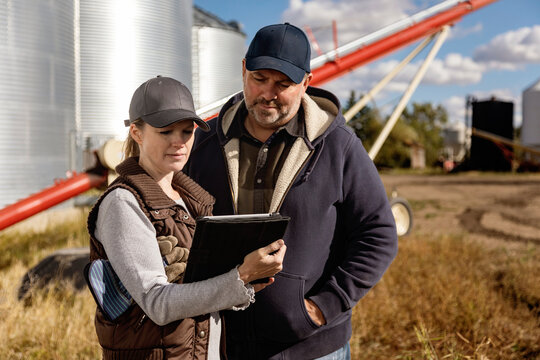 Mature couple working on their farm, standing in front of grain bins and auger and consulting their tablet computer; Alcomdale, Alberta, Canada