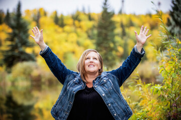 A woman enjoying quality time outdoors and taking time to raise her hands in worship at a city park with a lake in the background; Edmonton, Alberta, Canada