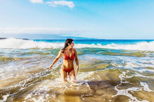 A woman in a red bikini stands in the ocean water off the shore at D. T. Fleming Beach with the Island of Molokai in the distance; Kapalua, Maui, Hawaii, United States of America