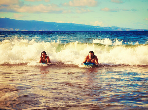 Happy married couple playing in the waves with bodyboards at D. T. Fleming Beach, with the Island of Molokai in the background; Kapalua, Maui, Hawaii, United States of America