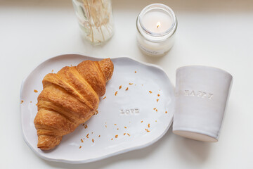 White glass and a plate on which there is a fresh croissant made of clay with the inscription love, handmade.