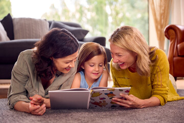 Same Sex Family With Two Mums And Daughter Lying On Floor Reading Book At Home Together