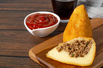 coxinha traditional brazilian snack fried in oil and stuffed with bovine meat or chicken meat,with one cutted in half. also with ketchup and soda on the background over wooden table.