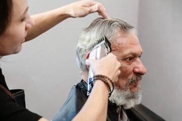 Working with hair clipper. Young professional barber girl cutting hair of mature bearded man. He is getting haircut at barbershop. Barbershop, beauty salon.