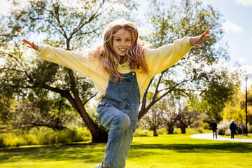 Young girl with long blond hair running across the grass and jumping towards the camera during a warm fall day in a city park; St. Albert, Alberta, Canada