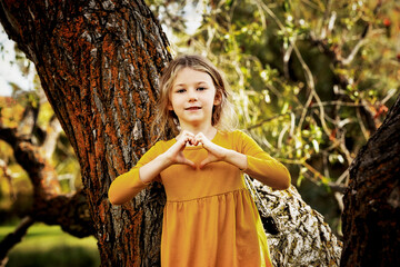 Young girl playing near a large tree and pausing for a picture while making a heart shape of love with her hands in a city park on a warm fall afternoon; St. Albert, Alberta, Canada