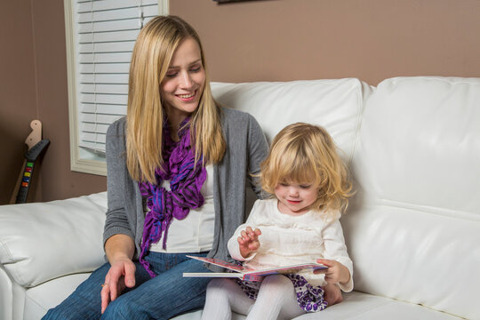 Young disabled mother sits on the couch at home reading to her toddler daughter; Spruce Grove, Alberta, Canada