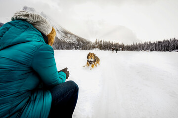 Woman calling her dog while hiking across frozen Emerald Lake with the Rocky Mountains in the background during winter in Yoho National Park; British Columbia, Canada
