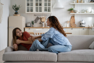 Happy positive young mother enjoying day with teenage girl at home, mom spending leisure carefree time with daughter at home, sitting on sofa together. Smiling mommy tickling laughing teen kid - obrazy, fototapety, plakaty