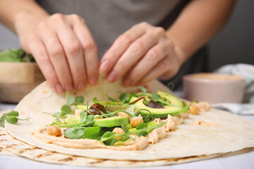 Woman making delicious hummus wrap with vegetables at table, closeup