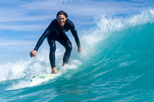 In water shot of a surfer on a wave surfing and smiling at the camera. Blue skies on sunny day with turquoise water. Surfer in black wetsuit on white board.