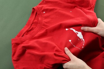 Woman holding red shirt with white paint stain on green background, top view