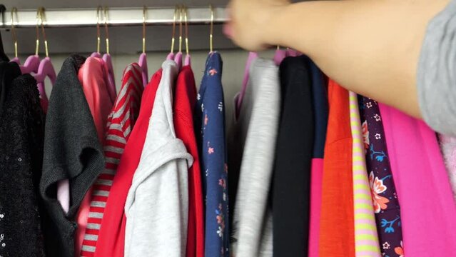 A lot of colorful women's clothes on pink hangers in the closet and a woman's hand moving clothes. Choice of clothes, order in the closet.