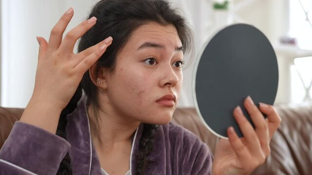 Close-up Asian young woman examining acne and wrinkles looking in hand mirror sitting indoors. Portrait of sad millennial lady with problematic skin at home in the morning