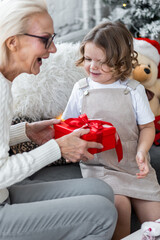 Attractive senior grandmother exchanging Christmas and New Year gift box with her granddaughter, cute little girl. Both looking happy and excited sitting near decorated tree in red Santa hats