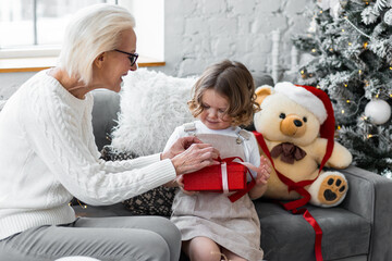 Attractive senior grandmother exchanging Christmas and New Year gift box with her granddaughter, cute little girl. Both looking happy and excited sitting near decorated tree in red Santa hats