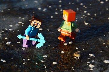 Fototapeta premium LEGO Minecraft figures of Steve and Alex trying to break thick layer of real ice on a lake with diamond pickaxe.
