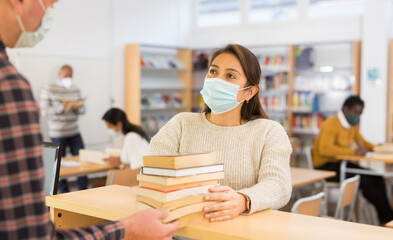 Young hispanic woman in protective face mask talking to librarian while returning books to...