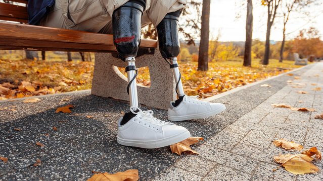 View of a man with prosthetic legs and white sneakers. Sitting on a bench in a park. Fallen yellow leaves around