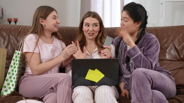 Front view three cheerful beautiful women talking laughing shopping online on pajama party. Caucasian and Asian millennial friends enjoying leisure choosing Black Friday purchases chatting