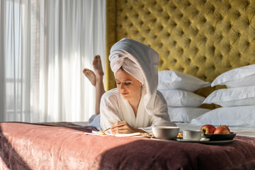 Pleasant domestic business woman in towel robe taking notes reminder in notepad lying on luxury bed