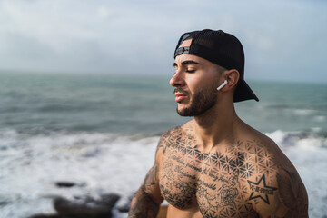 Chico joven musculado y tatuado posando en acantilado de una playa sin camiseta