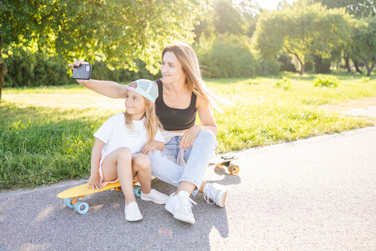 Woman with child sitting on skateboards in park and take photo by smartphone. Active family weekend.