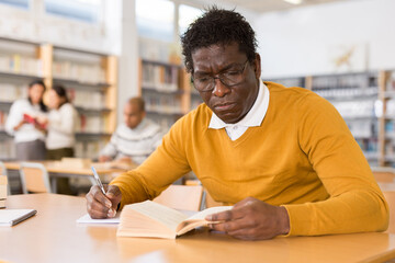 Obraz premium Intelligent adult aframerican man spending time in library, reading books and making notes. Self-education concept..