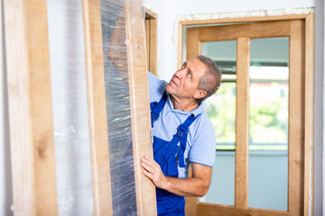 Serious male carpenter in uniform holding a wooden door for installing indoors