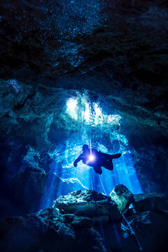 A cave diver descends into a cave system with light rays penetrating the water in the background.