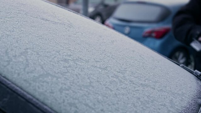 A man in a winter coat uses an ice scraper to remove heavy frost and ice from the windows and windshield of a frozen car on a sub-zero Winter morning.
