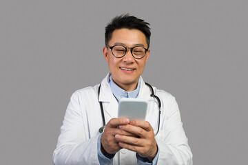 Cheerful adult japanese male therapist in white coat and glasses with stethoscope typing on phone