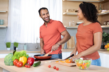 Smiling young black husband and wife in red t-shirts cuts salad, prepares lunch, cooking together in kitchen