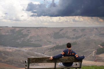 Young man sitting alone on a bench overlooking the mountain and valley. Thinking about life. Spending time alone and thinking about nature. Relaxed and peaceful atmosphere. 