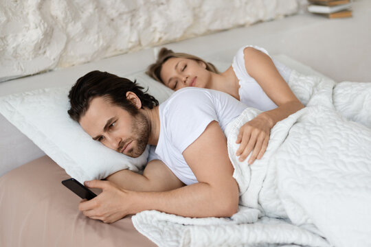 Angry man checking his sleeping girlfriend's smartphone, lying in bed at home, husband feeling jealous
