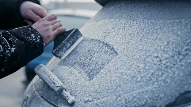 A man in a winter coat uses an ice scraper to remove heavy frost and ice from the windows and windshield of a frozen car on a sub-zero Winter morning.