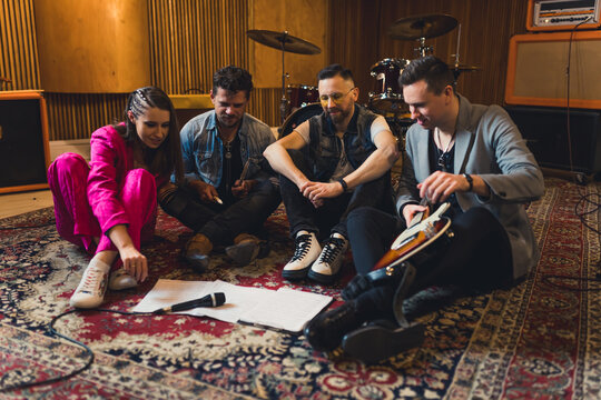 Full-length indoor view of a vocalist and three other musicians sitting on colourful carpet and writing music. Recording studio interior. High quality photo