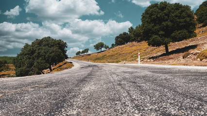 View of sky, trees, maquis, and clouds along a worn asphalt road in an Aegean town. Nature and sky landscape on a weathered asphalt mountain road