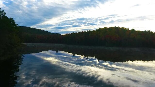Drone flying over stunning pond reflection blue sky and clouds and surrounded by mountains of fall foliage in morning light