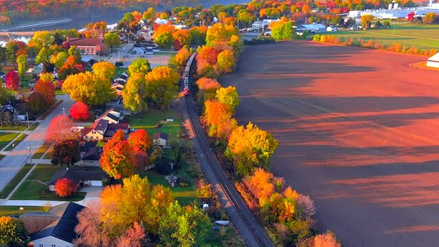 Freight train rolls through small town, all ablaze with Fall colors, aerial view.
