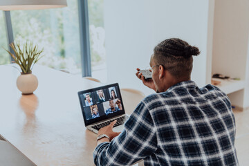 Fototapeta na wymiar African American man in glasses sitting at a table in a modern living room, using a laptop and smartphone for business video chat, conversation with friends and entertainment