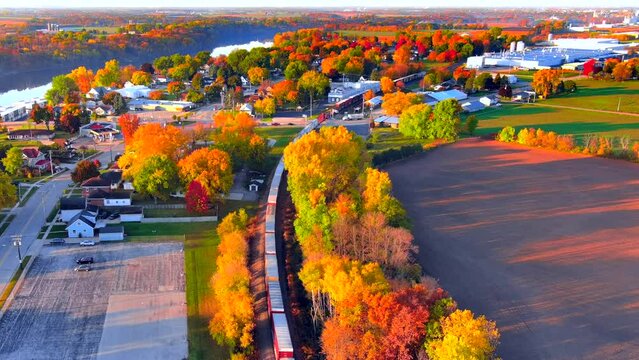 Freight train rolls through small town, all ablaze with Fall colors, aerial view.
